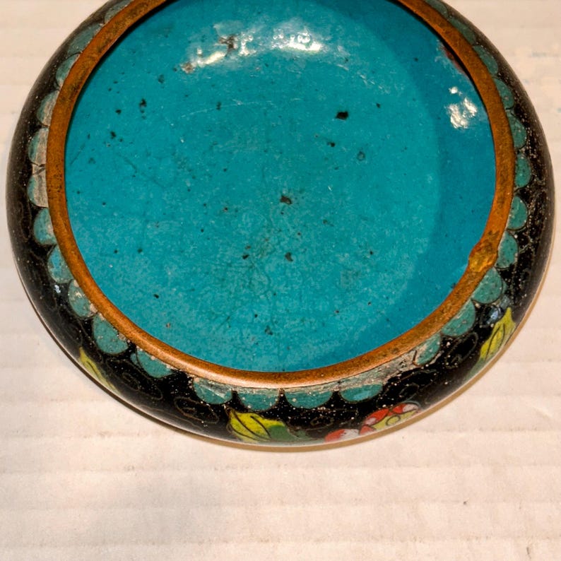 K&ouml;nnte beinhalten: Eine kleine, runde, antike Cloisonn&eacute;-Schale mit leuchtend t&uuml;rkisfarbenem Innenraum. Die Schale hat eine schwarze Au&szlig;enseite mit einem dekorativen Rand aus Blumen- und Blattmustern in verschiedenen Farben. Ein d&uuml;nnes, goldfarbenes Metallband umrandet den Rand.