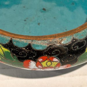 K&ouml;nnte beinhalten: Nahaufnahme einer dekorativen, antiken Cloisonn&eacute;-Schale. Die Schale hat einen t&uuml;rkisfarbenen Innenraum und ein detailliertes Blumendesign mit roten, gelben und wei&szlig;en Blumen auf schwarzem Hintergrund. Der Rand ist kupferfarben.