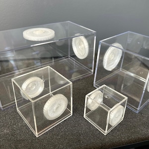 May include: A collection of clear acrylic display boxes in various sizes. Each box features a circular, textured, white accent on the sides and top. The boxes are arranged on a dark gray surface.