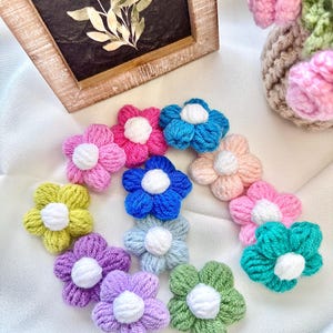 May include: A collection of colorful, crocheted flower appliques in various shades of pink, blue, green, yellow, and purple, each with a white center. The flowers are arranged on a white surface, with a framed botanical print in the background.