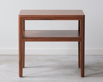 May include: A rectangular wooden side table with a shelf. The table is made of dark wood with a natural grain pattern. The table has a simple design with straight lines and a rectangular shape.