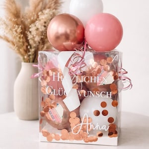 Könnte beinhalten: Eine transparente Geschenkbox gefüllt mit rosa und roségoldenen Luftballons, Konfetti und einem weißen Band. Auf der Box steht in weißer Schrift "Herzlichen Glückwunsch Anna". Im Hintergrund eine Vase mit Trockenblumen.