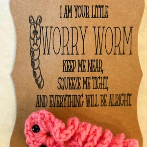 Puede incluir: Un gusano de preocupación rosa tejido a ganchillo con ojos de cuentas negras, sujeto a una tarjeta marrón. La tarjeta dice: "SOY TU PEQUEÑO GUSANO DE PREOCUPACIÓN, MANTENME CERCA, APRIÉTAME FUERTE, Y TODO ESTARÁ BIEN".