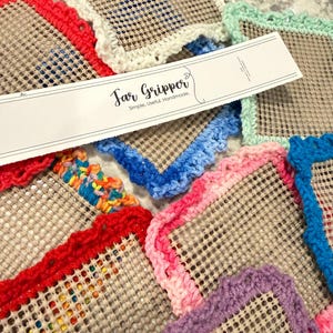 Peut inclure: Plusieurs dessous de bocal faits à la main et colorés. Chaque dessous de bocal a une base carrée en filet avec une bordure crochetée de différentes couleurs, notamment rouge, bleu, rose et violet. Le texte "Jar Gripper" est visible sur une étiquette blanche.