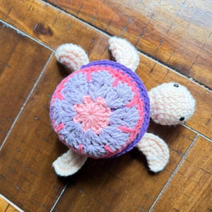 Puede incluir: Un juguete de tortuga tejido a ganchillo con cabeza y cuatro aletas de color melocotón claro. El caparazón presenta un patrón circular en tonos rosa, lavanda y morado oscuro, con dos pequeñas cuentas negras como ojos en la cabeza.