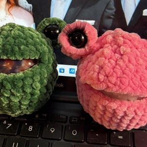 Può includere: Due figure rotonde all'uncinetto simili a rane. Una è verde scuro con occhi neri, l'altra è rosa con occhi neri e lunghe ciglia scure. Entrambe presentano ampie aperture orizzontali al centro del corpo che rivelano il contenuto interno.