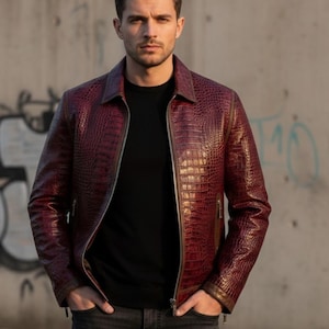 Könnte beinhalten: Burgunderfarbene Lederjacke mit Krokodilhautstruktur. Die Jacke hat einen Reißverschluss, einen Kragen und zwei Seitentaschen. Das Model trägt ein schwarzes Hemd und dunkelgraue Jeans.