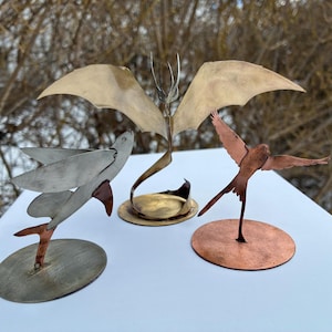 Könnte beinhalten: Drei Metallvogel-Skulpturen. Eine ist silberfarben mit Kupferakzenten, eine andere ist bronze mit ausgebreiteten Flügeln, und die dritte ist ein stilisierter Vogel mit großen Flügeln. Jede Skulptur ist auf einem kreisförmigen Sockel montiert.