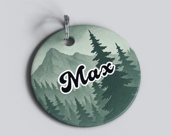 May include: A round pet tag with a forest scene in shades of green and gray. The tag features a mountain range and evergreen trees. The name "Max" is written in a black script font with a white outline.