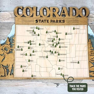 May include: Wooden Colorado State Parks map with laser-cut details. The map features a light wood finish with the state outline and park locations marked with small tree icons. The text "COLORADO STATE PARKS" is at the top, with the text "TRACK THE PARKS YOU VISITED" at the bottom.