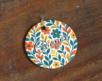 May include: Round floral tag with the name "Cooper" in black script. The tag features a cream background with orange, teal, and yellow flowers and green leaves. A clear plastic loop is attached at the top.