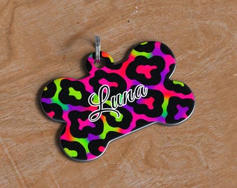 May include: A bone-shaped pet tag featuring a neon leopard print in pink, green, purple, and black. The name "Luna" is written in white script. A silver metal ring is attached at the top for easy attachment to a collar.