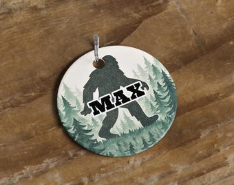 May include: A round, white ceramic ornament featuring a silhouette of Bigfoot walking through a forest of green trees. The word "MAX" is printed across the Bigfoot in a bold, black font. A silver metal loop is attached at the top.