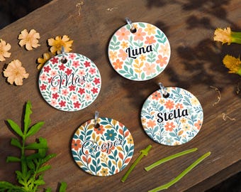 May include: Four round pet tags with floral designs and names: Max, Luna, Stella, and Cooper. Each tag has a silver-colored ring for attachment. The tags are set on a wooden surface with scattered flowers and greenery.