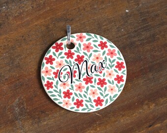 May include: A round, white floral tag with the name "Max" in black script. The tag features a pattern of red and pink flowers with green leaves. A silver metal hook is attached at the top.