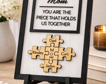 May include: A framed wooden sign with the words "Mom, You are the piece that holds us together." A wooden puzzle with the names Dad, Kinsley, Curt, Jadyn, and Mom is below the text. The frame is black.