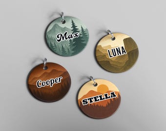 May include: Four round pet tags with mountain and forest designs. Each tag features a different name: Max, Luna, Cooper, and Stella. The tags have a silver-colored loop at the top for attaching to a collar. The color palette includes greens, browns, and oranges.