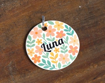 May include: A round, white pet tag with a floral pattern in peach, yellow, and green. The name "Luna" is written in black script across the center. A silver metal loop is at the top for attachment.