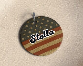 May include: A round wooden tag with an American flag design. The top half features gold stars on a dark green background, and the bottom half has red and brown stripes. The name "Stella" is written in a white, cursive font with a black outline.