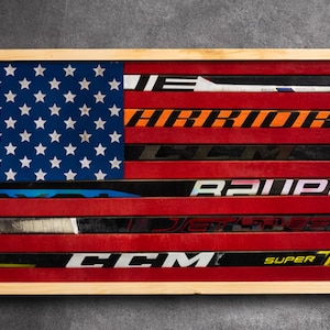 May include: Wooden framed artwork depicting the American flag. The flag's stripes are made from red hockey sticks, with a blue field of white stars in the upper left corner. The frame is made of light-colored wood.