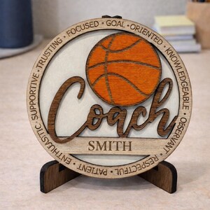 May include: A wooden basketball coach plaque with the word "Coach" in a cursive font, and the name "SMITH" below. The plaque features a basketball graphic and words like "Focused" and "Supportive" around the edge. It stands on a wooden base.