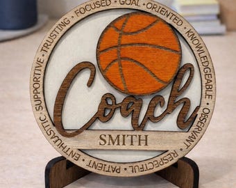 May include: A wooden basketball coach plaque with the word "Coach" in a cursive font, and the name "SMITH" below. The plaque features a basketball graphic and words like "Focused" and "Supportive" around the edge. It stands on a wooden base.