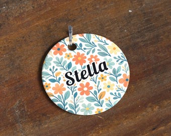 May include: A round, floral-patterned tag with the name "Stella" in black script. The tag features a white background with orange, yellow, and blue flowers and green leaves. A silver metal loop is attached at the top.
