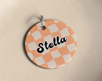 May include: A round, peach-colored tag with a white checkerboard pattern. The word "Stella" is written in black script across the center. A silver metal ring is attached at the top for hanging.