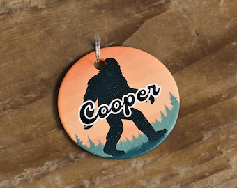 May include: Round Bigfoot-themed ornament with a silhouetted figure walking across a sunset background. The name "Cooper" is written in a cursive font across the figure. A silver-colored metal loop is at the top for hanging.