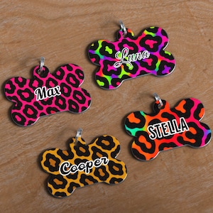 May include: Four dog tags in a bone shape with a leopard print design. Each tag has a different color scheme: pink, green, orange, and yellow. The names "Max", "Luna", "Stella", and "Cooper" are printed on each tag.