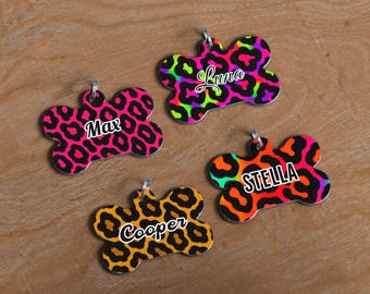 May include: Four dog tags in a bone shape with a leopard print design. Each tag has a different color scheme: pink, green, orange, and yellow. The names "Max", "Luna", "Stella", and "Cooper" are printed on each tag.