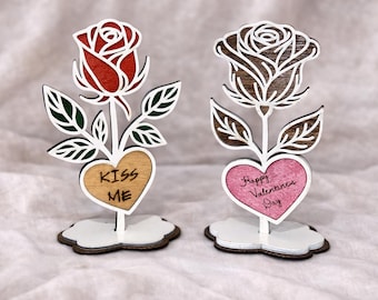 May include: Two wooden rose decorations with heart-shaped bases. One rose is red, the other is brown. The first heart says "KISS ME", the second says "Happy Valentine's Day". The leaves are green.