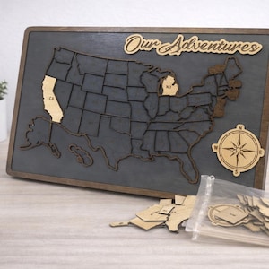 May include: Wooden map of the United States with the words "Our Adventures" in a cursive font. The map is gray with brown borders and includes wooden state cutouts. A compass and additional state cutouts are included.