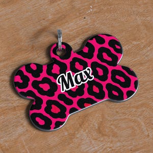 May include: A bone-shaped pet tag with a pink leopard print design and the name "Max" in white script. The tag has a silver-colored loop for attaching to a collar. The background is a light brown wooden surface.