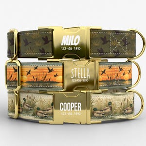 May include: Three dog collars with gold-tone metal buckles and hardware. The top collar is olive green with a duck pattern, the middle features a sunset marsh scene, and the bottom shows ducks in reeds. Each buckle is engraved with a name and phone number.
