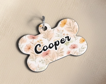 May include: Bone-shaped pet tag with a floral design in soft peach, yellow, and green hues. The name "Cooper" is written in a bold, black script across the center. A silver-colored metal ring is attached at the top.