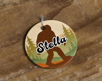 May include: A round, Bigfoot-themed tag with the name "Stella" in a whimsical font. The design features a brown Bigfoot silhouette walking through a forest scene with green trees and a tan background. A silver-colored metal loop is at the top.
