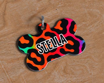May include: A bone-shaped pet tag with a vibrant leopard print design in neon orange, pink, black, and green. The name "STELLA" is displayed in bold white letters. The tag has a silver-colored ring for attachment.