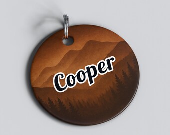 May include: A round, brown pet tag with a mountain and forest design. The name "Cooper" is written in white, outlined in black, across the center. A silver-colored ring is attached at the top for easy attachment to a collar.