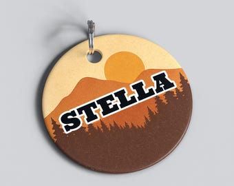 May include: A round pet tag with a mountain and sun design. The tag is tan with a brown bottom and a silhouetted forest. The word "STELLA" is in bold, black letters with a white outline.