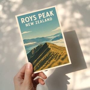 Op de afbeelding: Een ansichtkaart met een schilderachtig uitzicht op Roys Peak in Nieuw-Zeeland. De ansichtkaart heeft een blauw en beige kleurenschema, met de tekst "ROYS PEAK NEW ZEALAND" bovenaan. De afbeelding toont een bergketen met een pad.