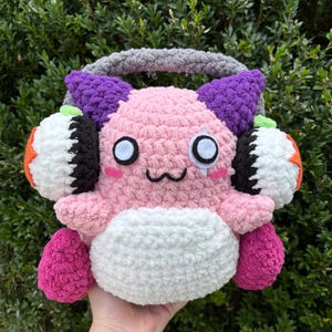 May include: A handmade, plush, pink creature with large white eyes, black accents, and a small smile. It has purple horns, orange and white ear coverings, and a grey handle. The creature is held against a backdrop of green foliage.