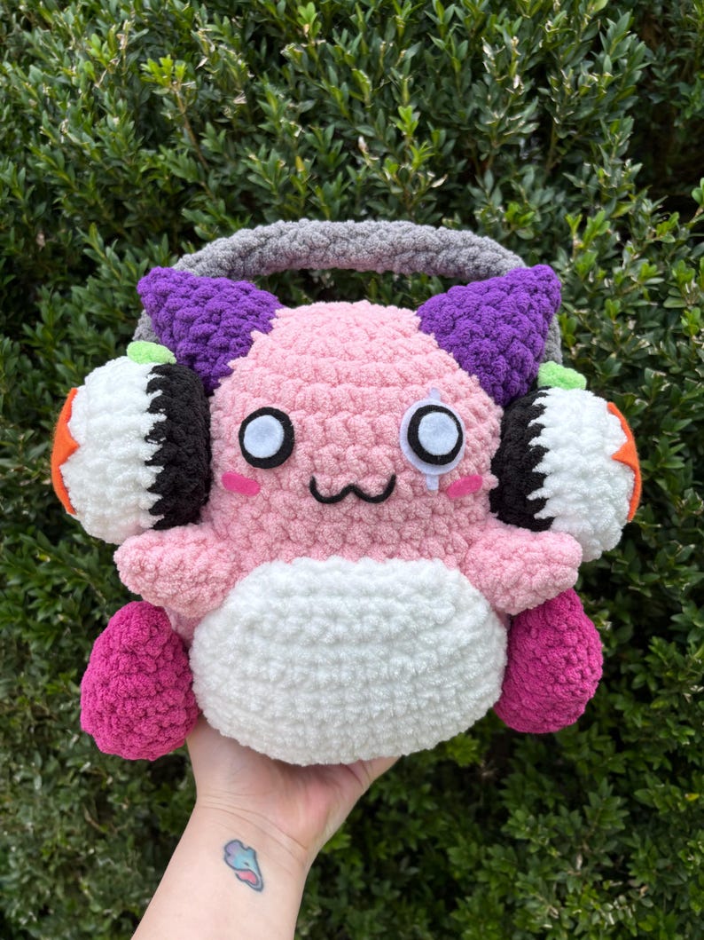 May include: A handmade, plush, pink creature with large white eyes, black accents, and a small smile. It has purple horns, orange and white ear coverings, and a grey handle. The creature is held against a backdrop of green foliage.