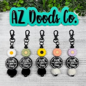 May include: Five keychains with black hardware, each featuring a different colored daisy, a black circle with the text "Faith Over Fear", and a fluffy pom-pom. The top of the image has the text "AZ Doods Co."