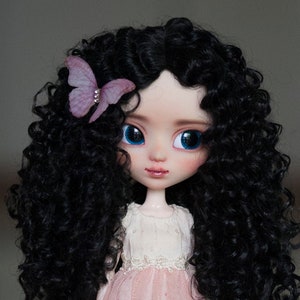 May include: A doll with large blue eyes and long, curly black hair. The doll wears a pink dress with a white lace trim and a butterfly hair accessory. The doll's face is delicately painted with rosy cheeks and a subtle lip color.