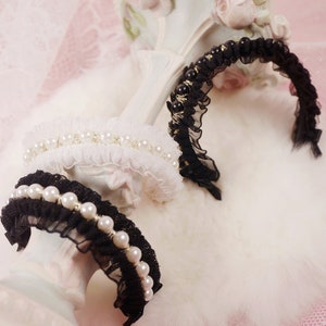 May include: Three decorative headbands in black and white, adorned with pearls and lace. The headbands are displayed on a white, fluffy surface, with a pink background and a decorative white stand.