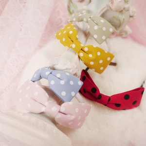 May include: A collection of colorful polka dot hair bows in pink, blue, yellow, and red. The bows are arranged on a white surface with a decorative, ornate object in the background. The bows are made of fabric and have a classic bow shape.
