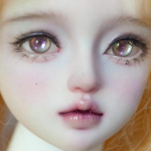 以下が含まれることがあります： 人形の顔のクローズアップ。肌は白く、頬はバラ色、唇はふっくらと艶やか。人形は紫色の虹彩と長い黒い睫毛を持つ大きな目をしている。波打つ金髪が顔を囲み、柔らかく幻想的な印象を与えている。