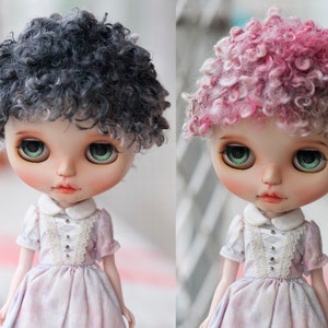 May include: Two dolls with different colored wigs. The doll on the left has a black and gray curly wig. The doll on the right has a pink and gray curly wig. Both dolls are wearing a white dress with lace trim.