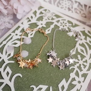 May include: Two delicate bracelets with star-shaped charms. One bracelet is gold with clear beads, the other is silver with clear beads. The bracelets are displayed on a green background with a white lace overlay.